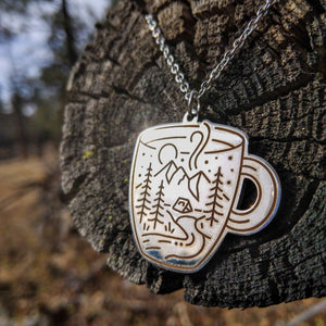 A Coffee cup-shaped pendant on a chain, with an etched scene of a tent amidst a mountain landscape, surrounded by trees and a stream, with a moon and stars in the background, made of stainless steel.