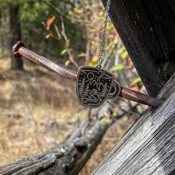 A Coffee cup-shaped pendant on a chain, with an etched scene of a tent amidst a mountain landscape, surrounded by trees and a stream, with a moon and stars in the background, made of stainless steel.