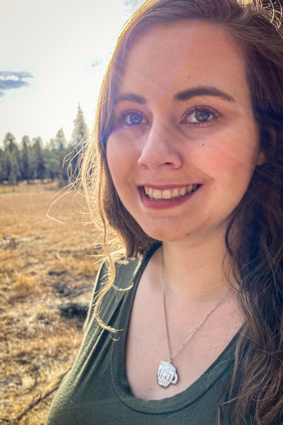 A girl outside wearing a Coffee cup-shaped pendant on a chain, with an etched scene of a tent amidst a mountain landscape, surrounded by trees and a stream, with a moon and stars in the background, made of stainless steel.